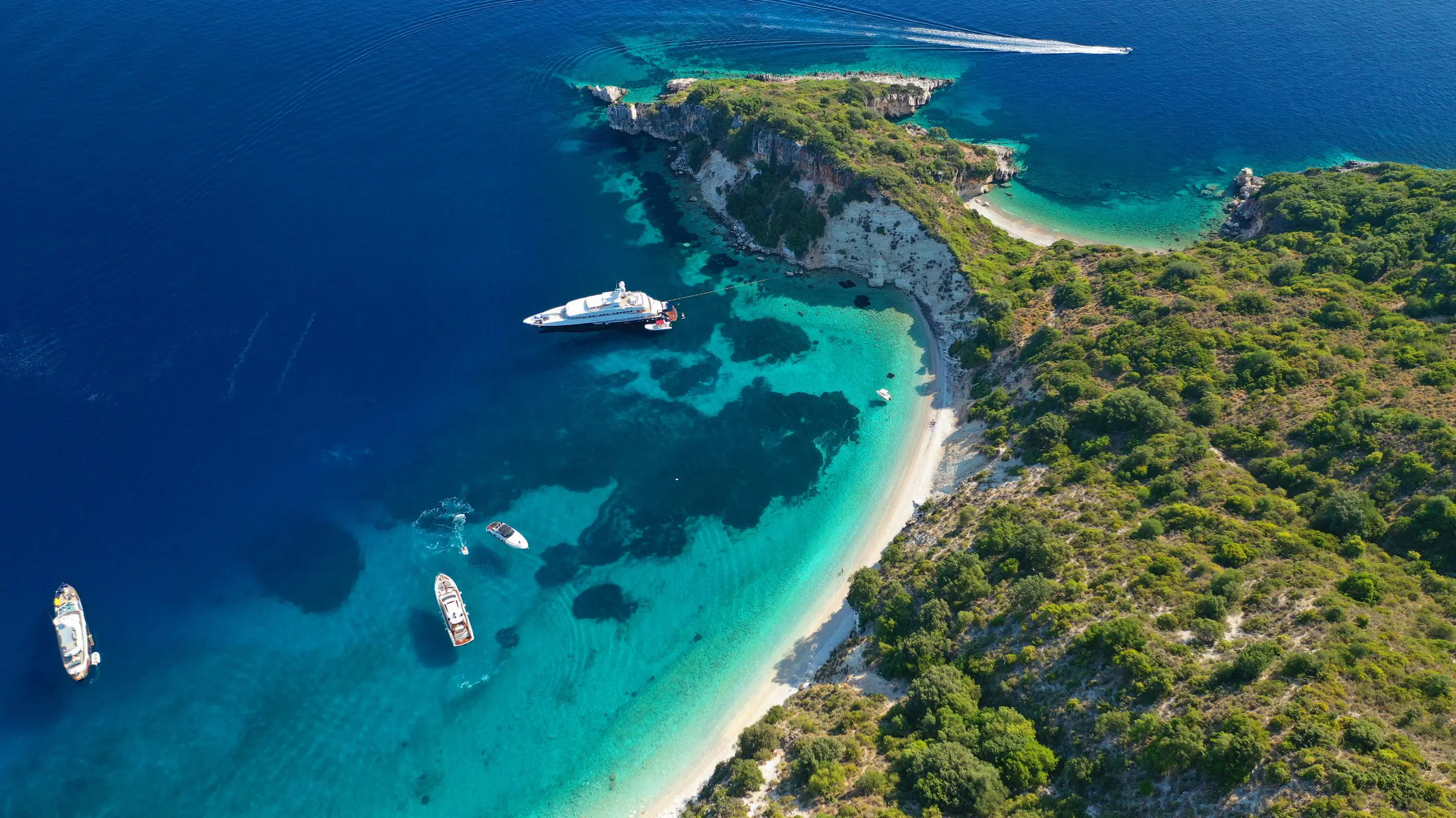 Bird's eye view of a sandy beach and bay of Gidaki, bright blue water with boats docked. Forested land in the right forefront