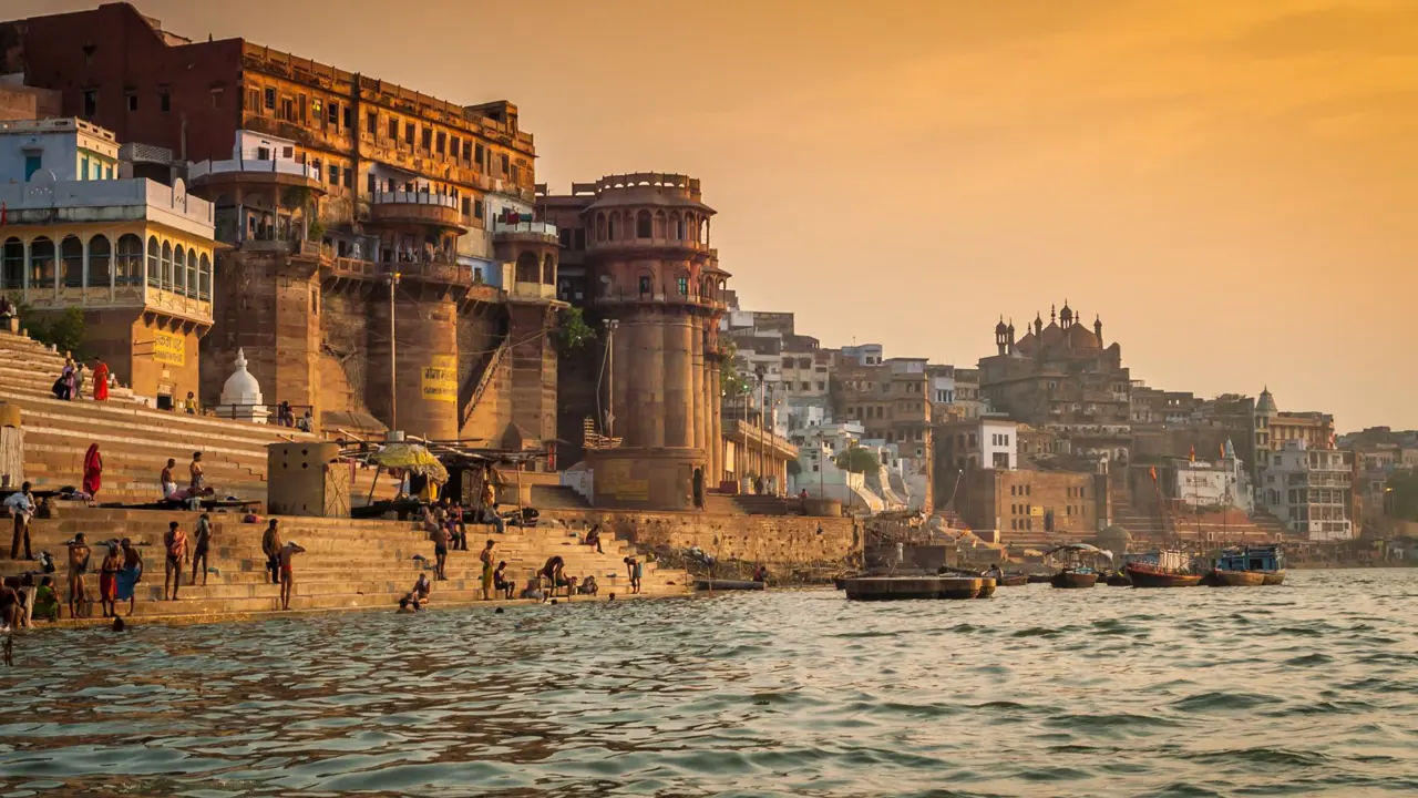 People gathering and bathing along the ghats of the River Ganges in Varanasi during sunset, with historic stone buildings and temples rising along the riverfront