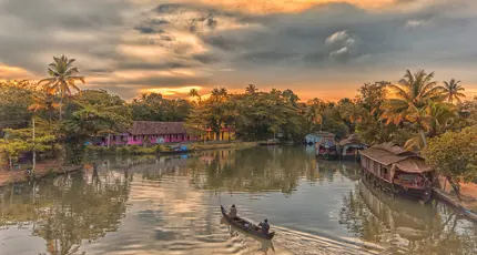 Two people in a canal boat on the Kerala Backwaters at sunset, with a pink house and palm trees lining the banks, and warm light reflecting on the calm water