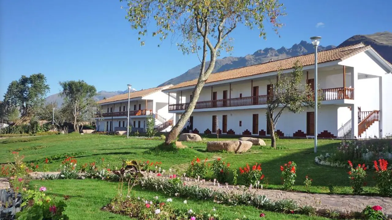 Exterior of Hotel Augustos in Urubamba, featuring a white colonial-style building with gardens, trees, and a mountain backdrop