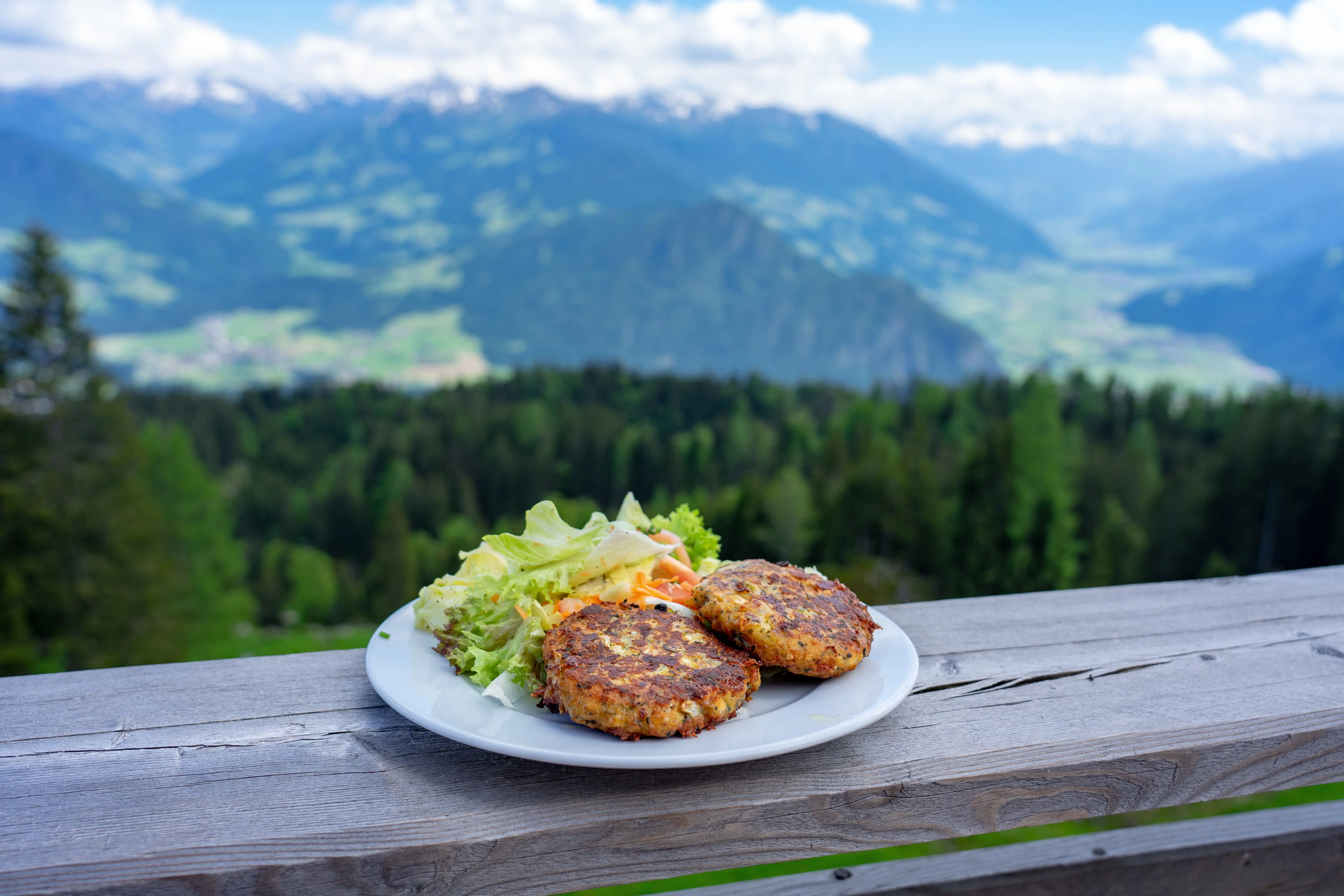 In-focus shot of a plate of flat, circular patties and a side salad on a wooden surface. Blurry, green mountains and trees in the background 