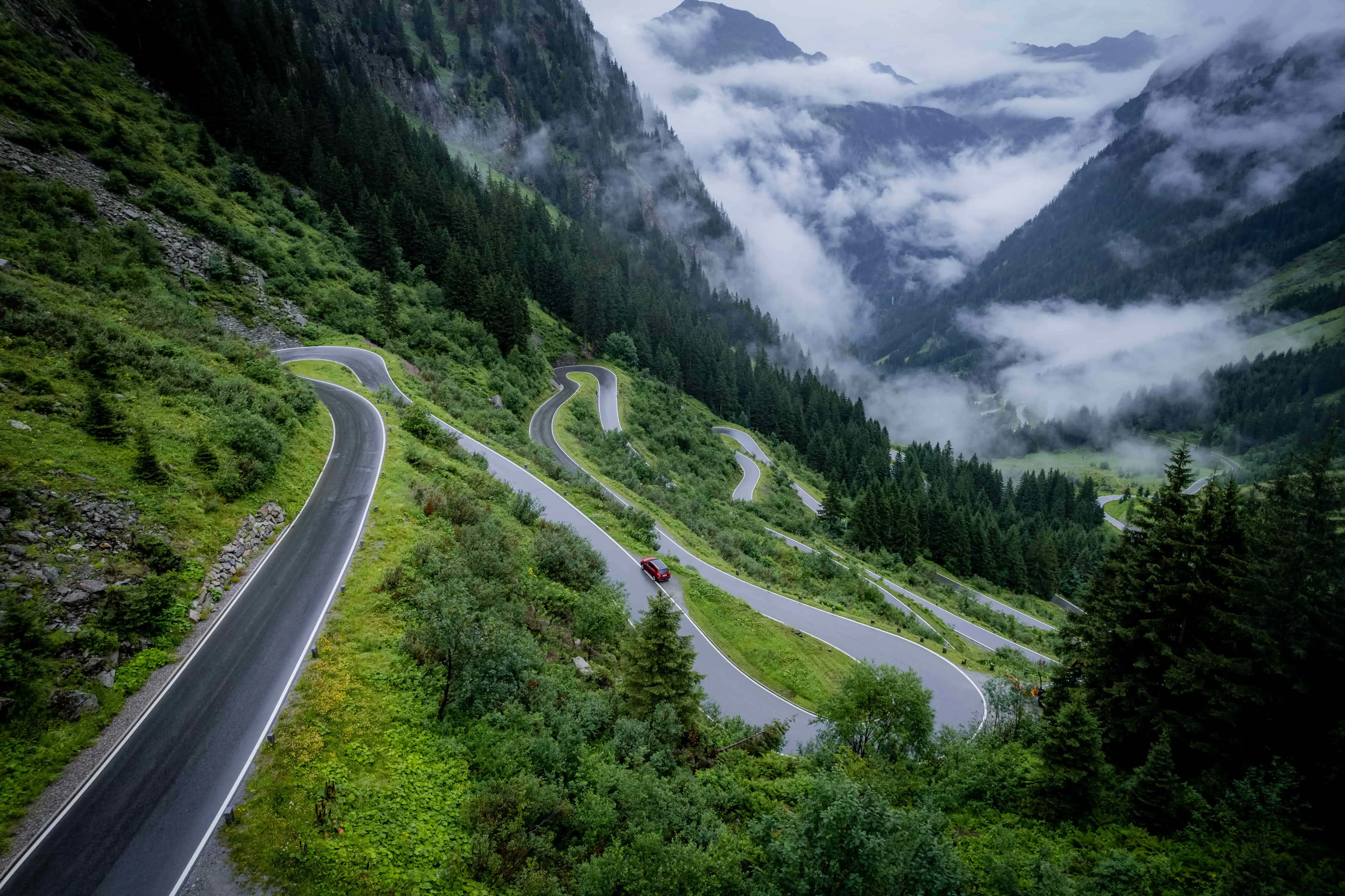 High angle shot of a road winding up a mountain in the Austrian Tyrol, with a red car on it. Fir trees cover the majority of the mountainsides, clouds further down the mountain and in the distance.