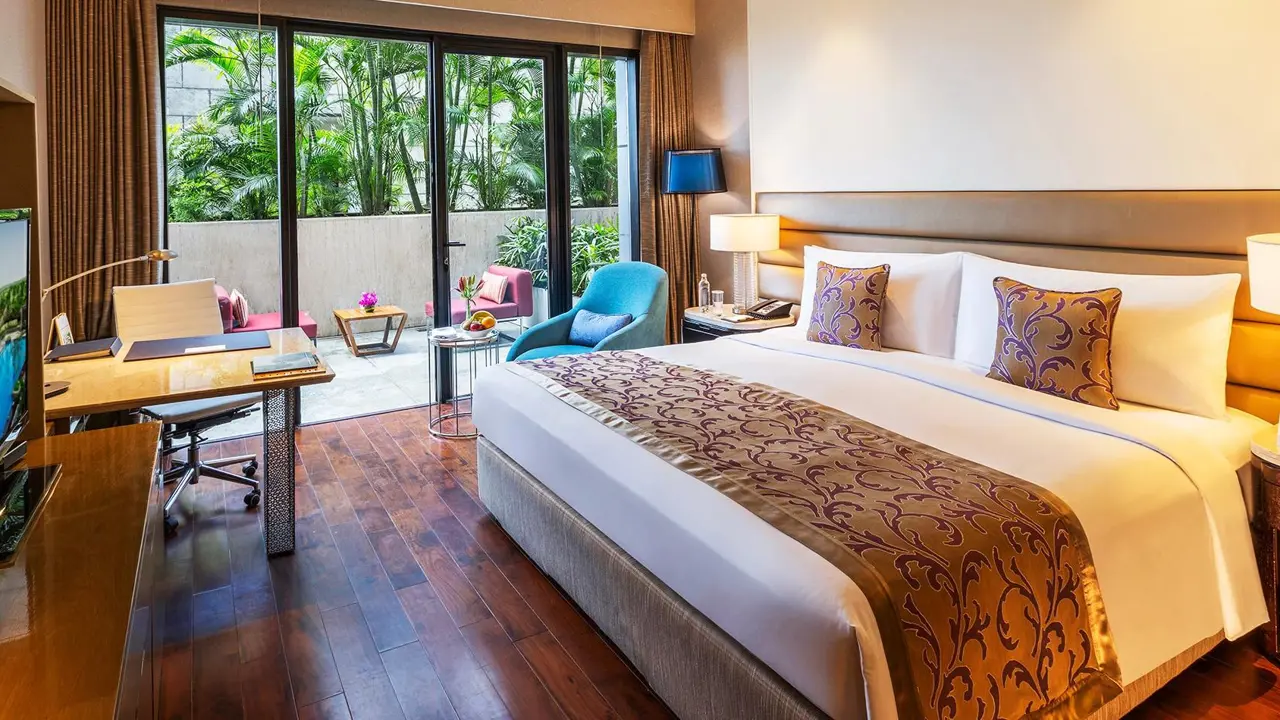Cosy double room at the Leela Ambience Convention Hotel in Delhi, featuring warm lighting, wooden floors, and a terrace outside the patio doors with two pink lounge chairs and a small coffee table