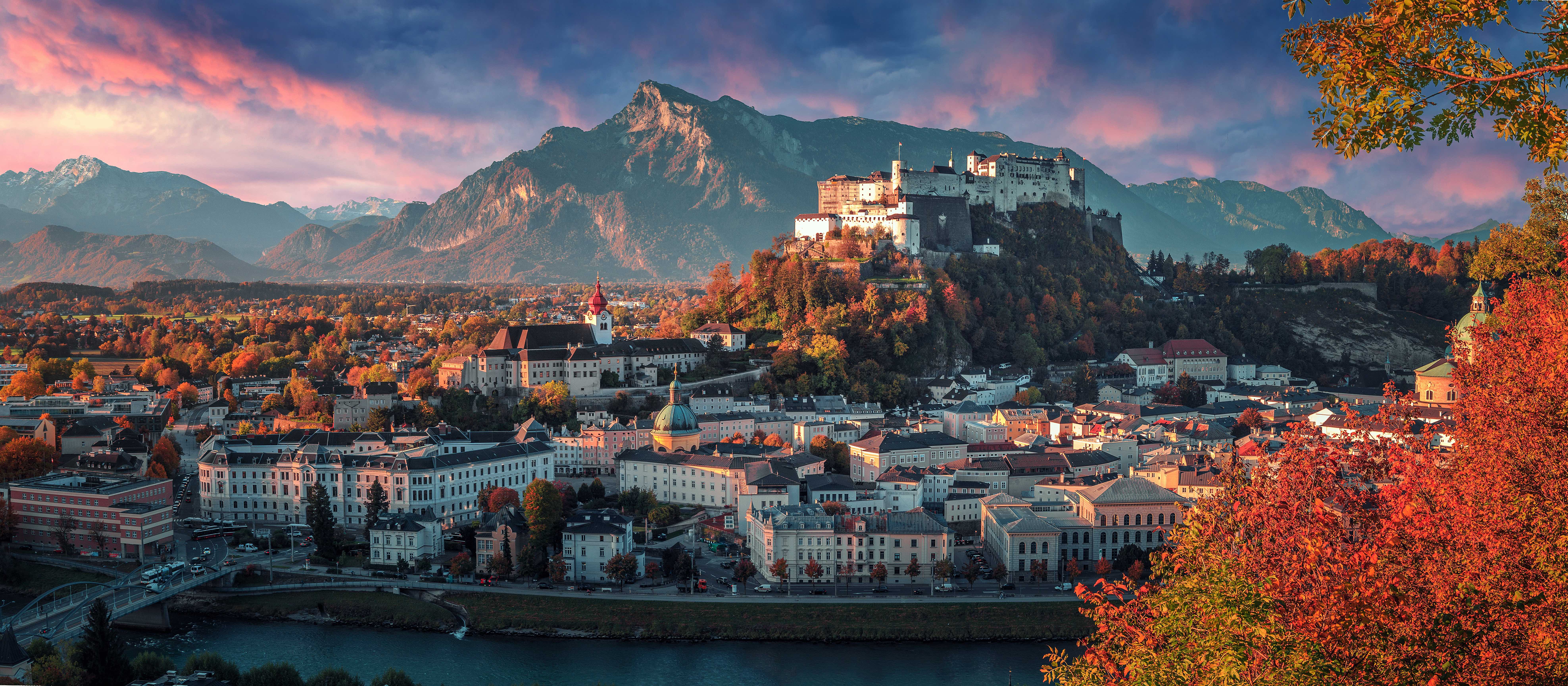 Wide angle shot of the city of Salzburg, showing the white and cream buildings, one with a turquoise turret and one further back with a red turret. Slightly to the right is a hill covered in green and orange trees with the fortress on the top. Mountains in the distance in front of blue and pink sky. In the right forefront, there is red and green tree leaves.