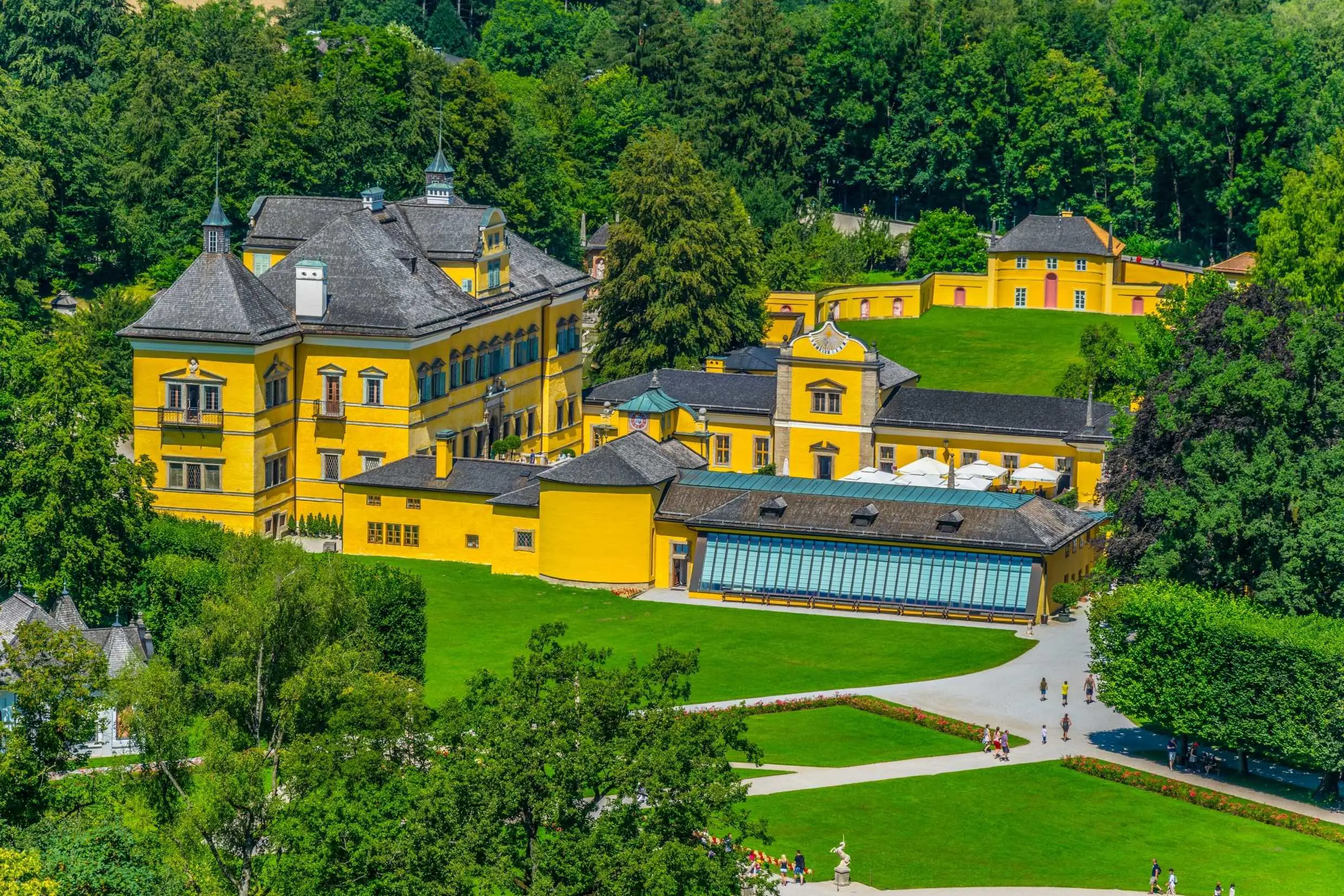Wide angle view of a yellow manor-like building with black roofs, with a green lawn out the front and trees surrounding it. In the forefront there are some more grassy lawns and pathways that go through them and up to the building, with people walking