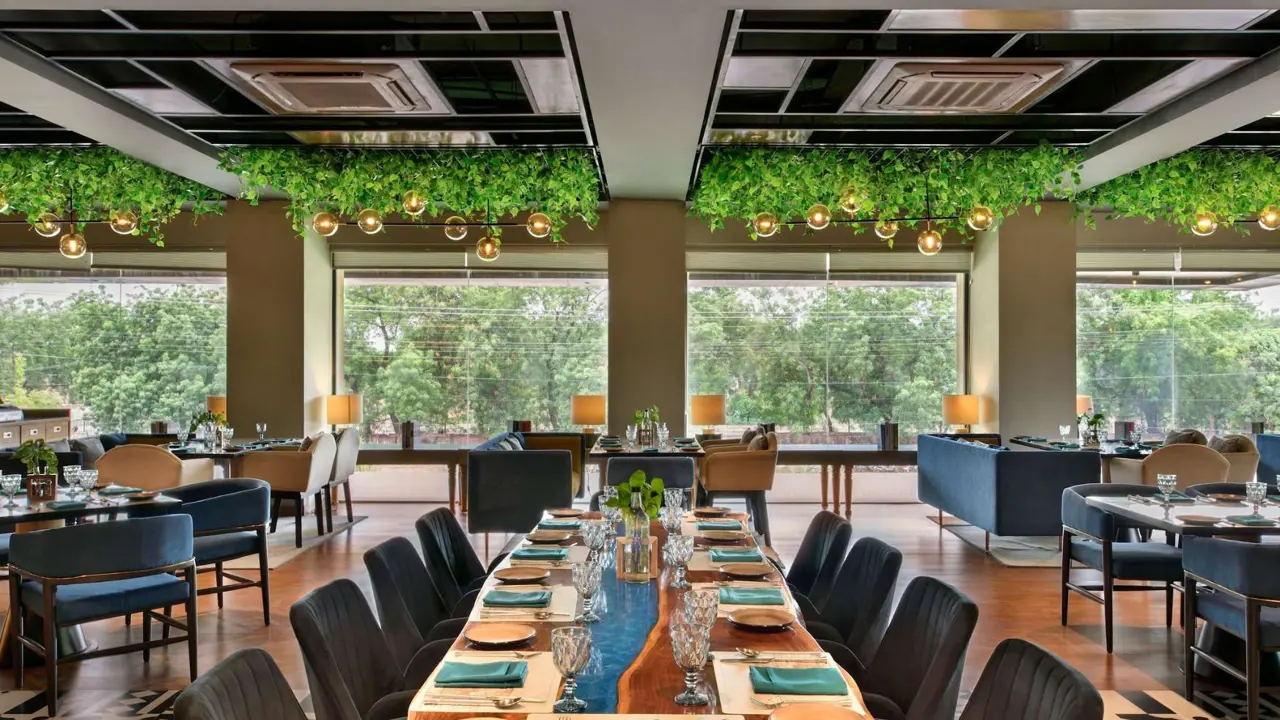 Modern restaurant at Novotel Jodhpur with large windows, hanging greenery, elegant table settings, and a mix of comfortable seating overlooking trees outside