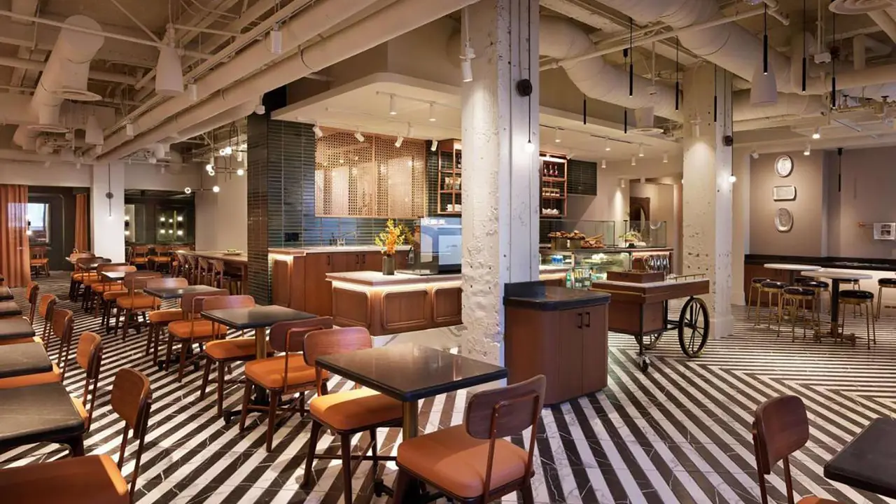 Interior of the stylish dining room at Clift Royal Sonesta, San Francisco, featuring black and white striped floors, an industrial-style white ceiling, dark wood tables, and seats with orange cushioning