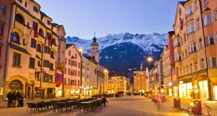 View of a wide road with shops and tall buildings either side. Tables and chairs are laid out in the road and bicycles are parked up on the right side. There is a clock tower which is taller than the rest of the buildings with a turquoise turret on the left, and snowy mountains can be seen at the end of the road, in front of light blue evening sky