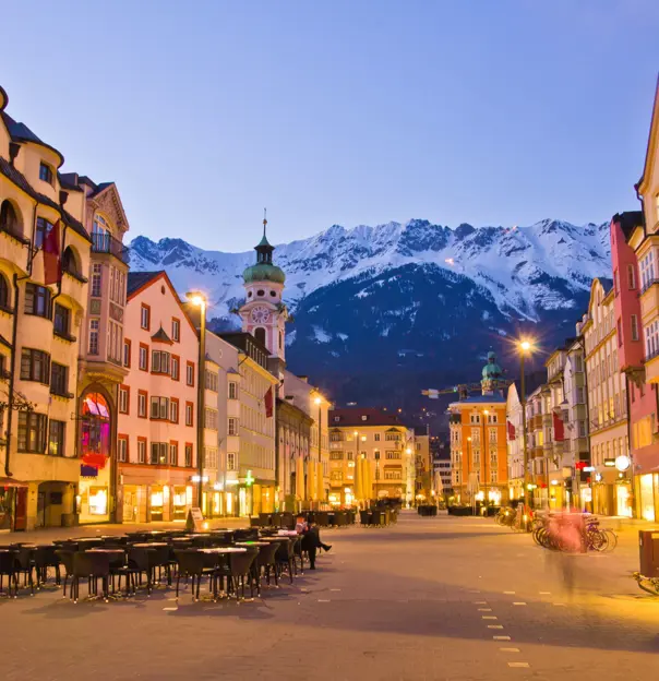 View of a wide road with shops and tall buildings either side. Tables and chairs are laid out in the road and bicycles are parked up on the right side. There is a clock tower which is taller than the rest of the buildings with a turquoise turret on the left, and snowy mountains can be seen at the end of the road, in front of light blue evening sky
