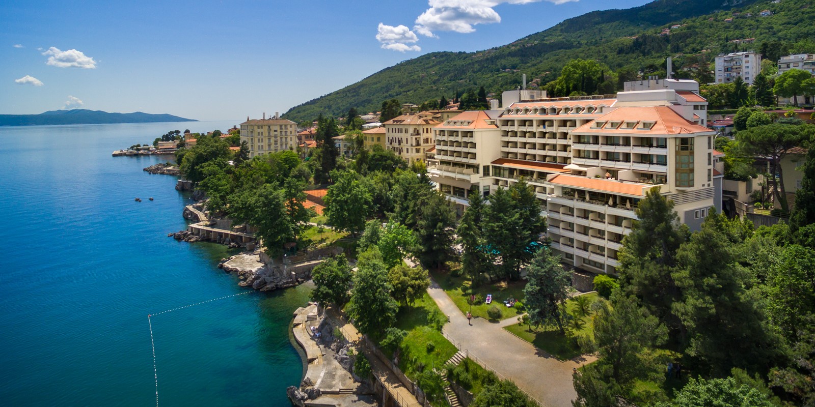Seaside view of Hotel Excelsior in Lovran, Croatia, with terraced buildings surrounded by greenery, overlooking the clear blue Adriatic Sea on a sunny day
