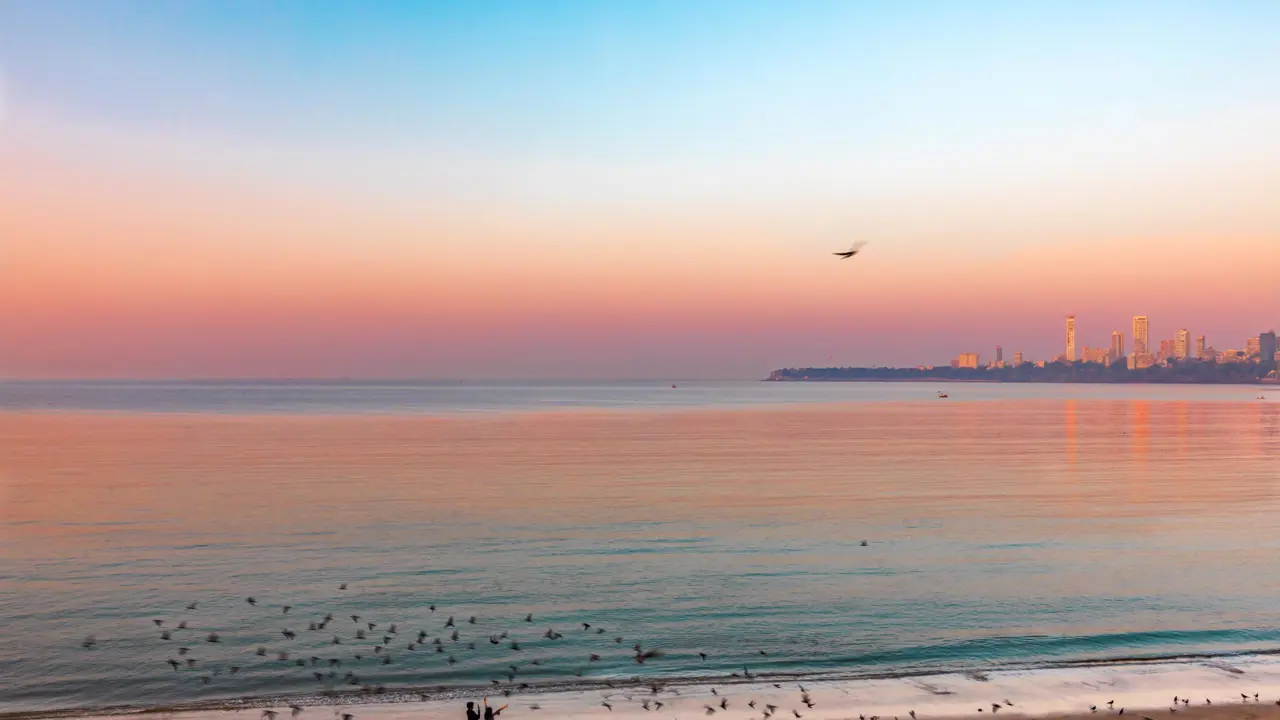 Mumbai seafront at sunset on a calm day, with birds flying over the water, two people standing on the beach, and the city skyline in the background