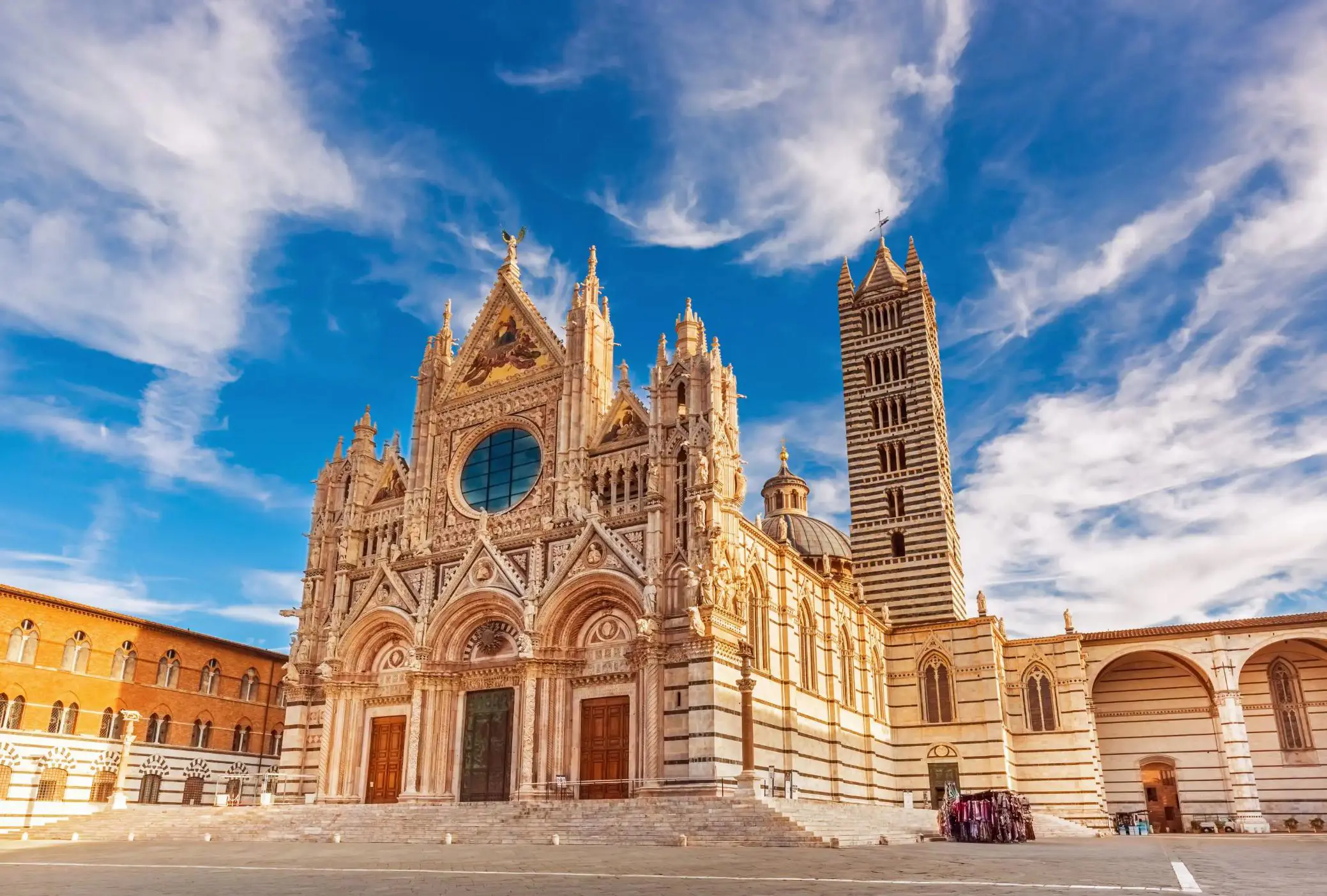 The intricate façade of Siena Cathedral in Italy, bathed in warm sunlight under a vivid blue sky with wispy clouds