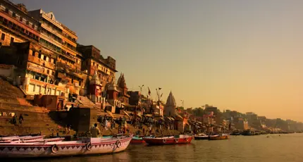 Boats anchored along the ghats of Varanasi at sunrise, with ancient sandstone buildings and temples glowing in golden light by the River Ganges