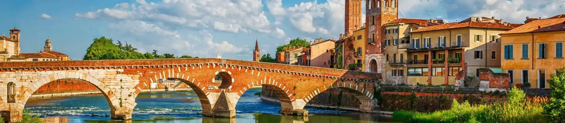 Scenic view of the historic Ponte Pietra bridge crossing the Adige River in Verona, Italy, with colourful old buildings and church towers under a bright blue sky with scattered clouds