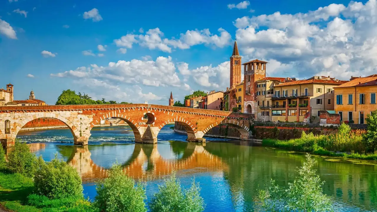 Scenic view of the historic Ponte Pietra bridge crossing the Adige River in Verona, Italy, with colourful old buildings and church towers under a bright blue sky with scattered clouds