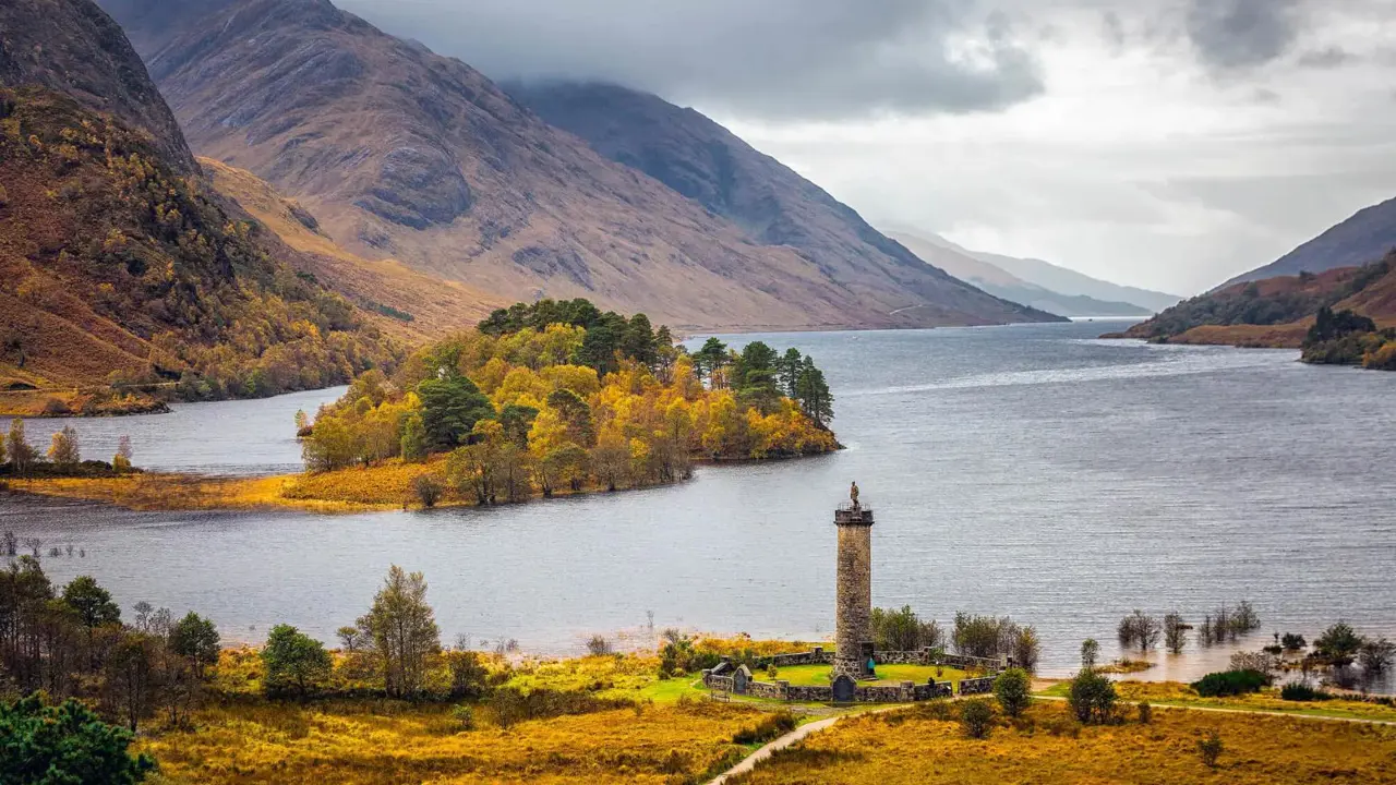 Glenfinnan Monument on a grassy hill at the head of Loch Shiel in the Scottish Highlands, with a lone statue of a Highlander atop the stone tower and dramatic mountains in the background