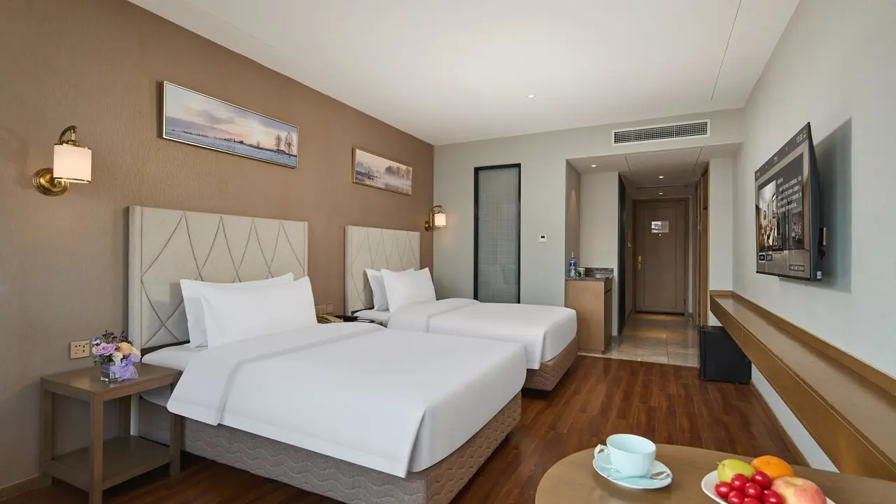 A contemporary hotel room at the Youngor Central Hotel in Suzhou, China, featuring two neatly made beds, a corner table with a fruit bowl, and a neutral yet inviting décor