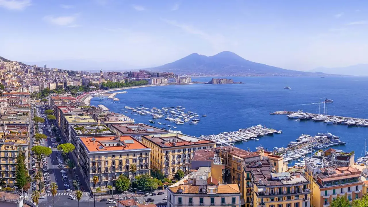 The Bay of Naples with Mount Vesuvius in the distance, lined with waterfront buildings and apartments, and a marina filled with boats