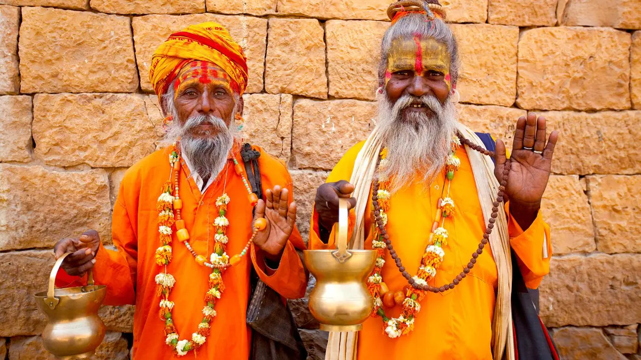 Two elderly Jain priests in orange robes and garlands stand before a sandstone wall, holding brass pots in Rajasthan in India