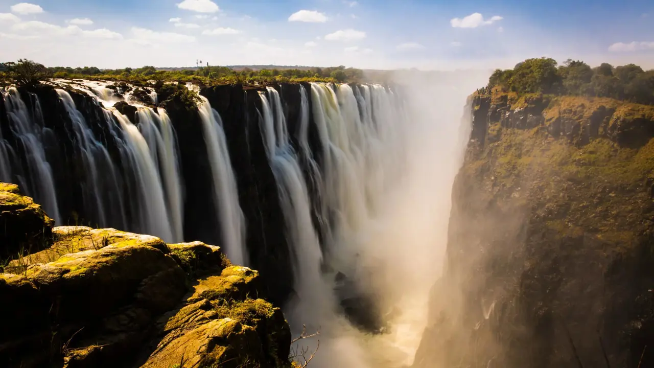Victoria Falls cascading powerfully over cliffs, surrounded by lush greenery under a bright blue sky with scattered clouds
