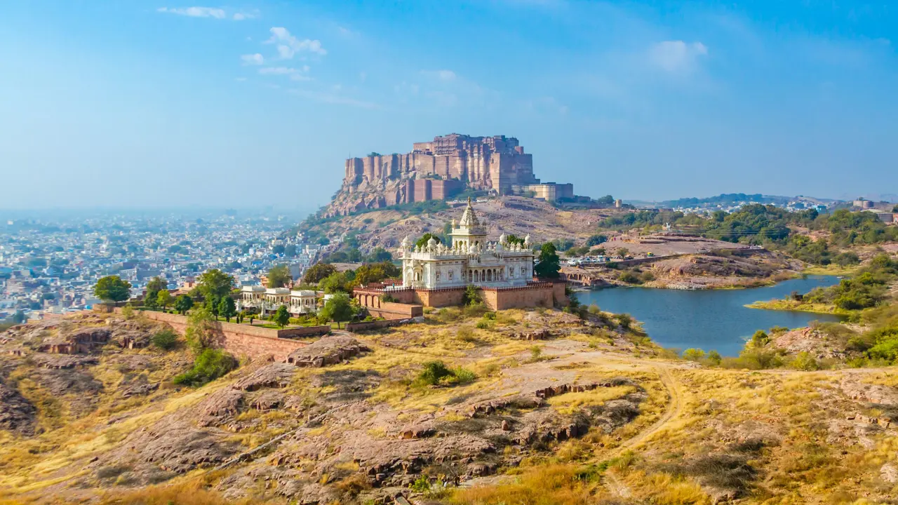 View of the white marble Jaswant Thada memorial and Mehrangarh Fort rising above Jodhpur, with a lake and cityscape in the background under a clear blue sky