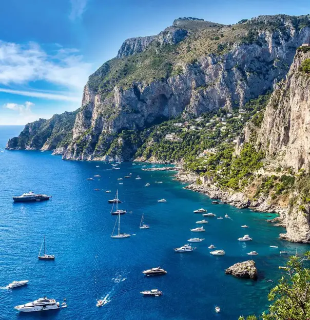 Speedboats and small yachts anchored in the turquoise waters beside Capri’s tree-covered cliffs