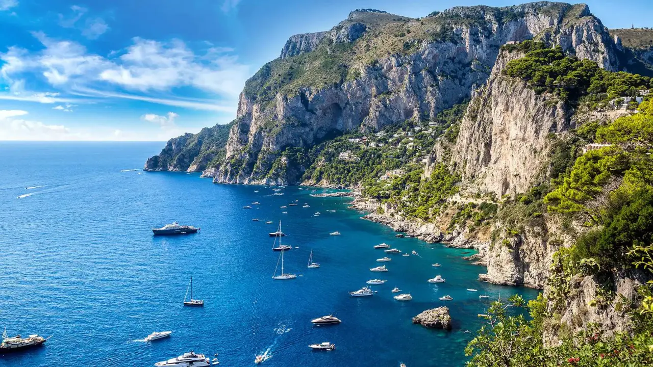 Speedboats and small yachts anchored in the turquoise waters beside Capri’s tree-covered cliffs