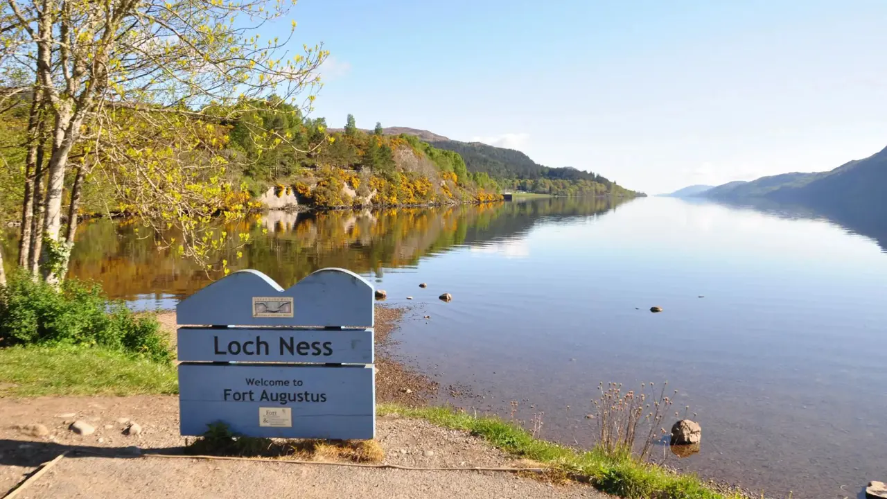 Calm waters of Loch Ness reflecting the surrounding hills under a clear blue sky, with a sign welcoming visitors to Fort Augustus on the shore