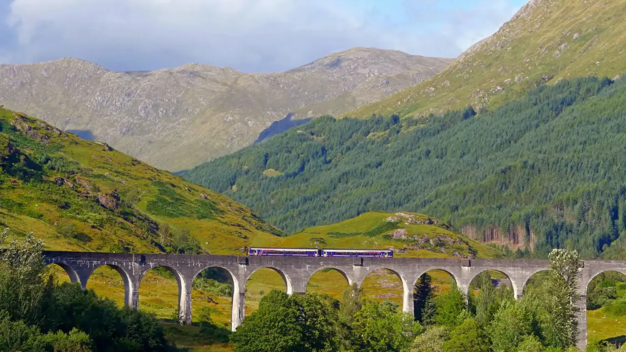 A scenic train travels across the Glenfinnan Viaduct in Scotland, surrounded by rolling green hills and forested mountains
