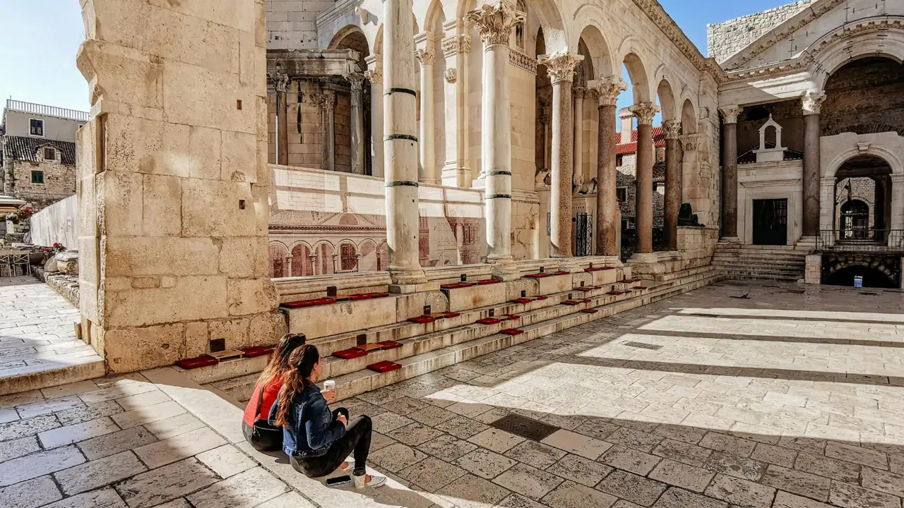 Two women sit on the iconic steps of the Peristyle courtyard in Diocletian’s Palace, surrounded by ancient Roman columns and white stone architecture