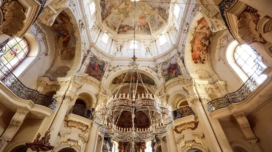 Under shot of a ceiling and chandelier, with old religious artwork and architectural design.