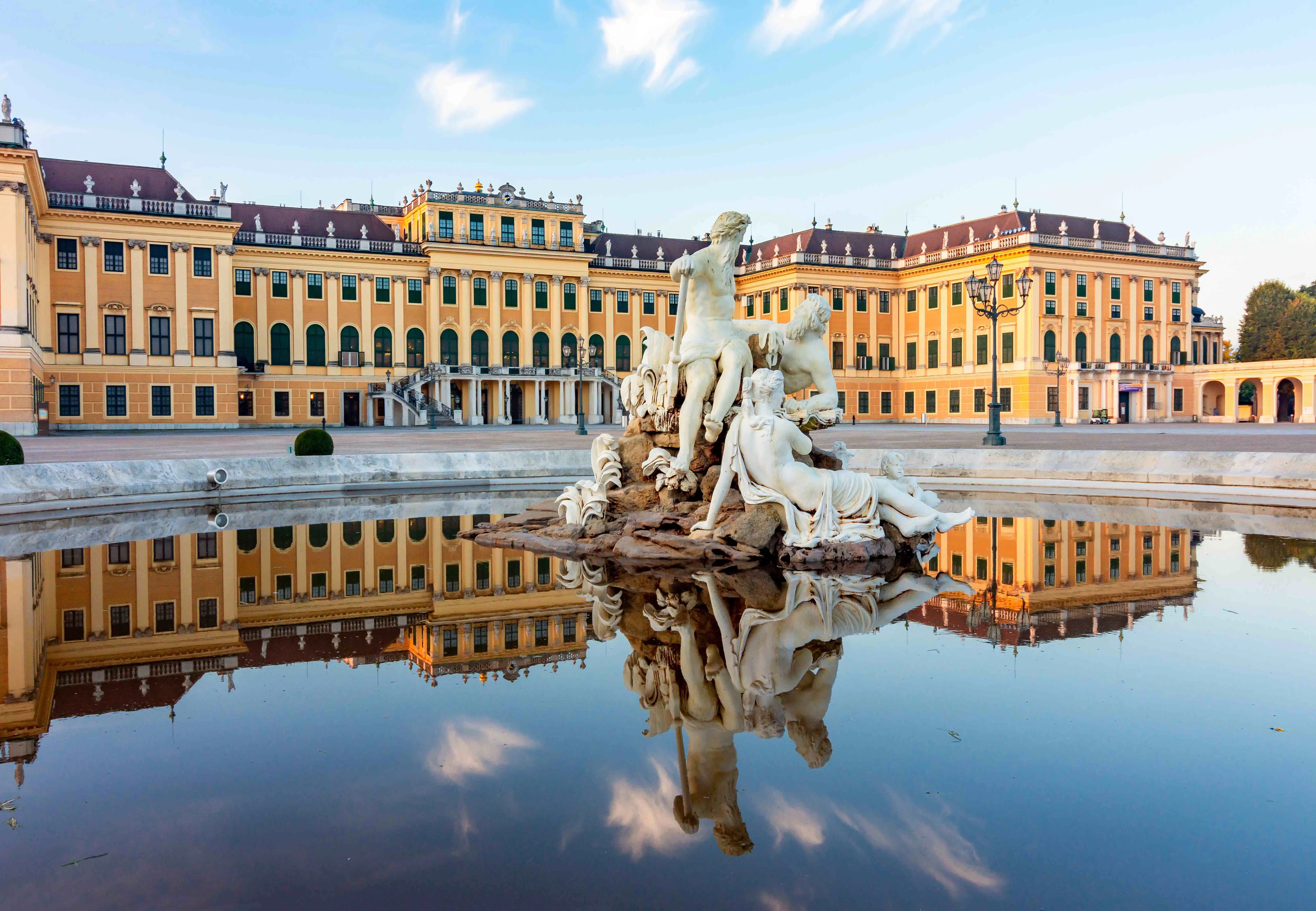 A fountain with a white statue of three people sitting on a rock, on top of water, where its reflection can be seen. Behind it is Schönbrunn Palace, yellow and beige wide building, in front of a blue sky with small clouds.