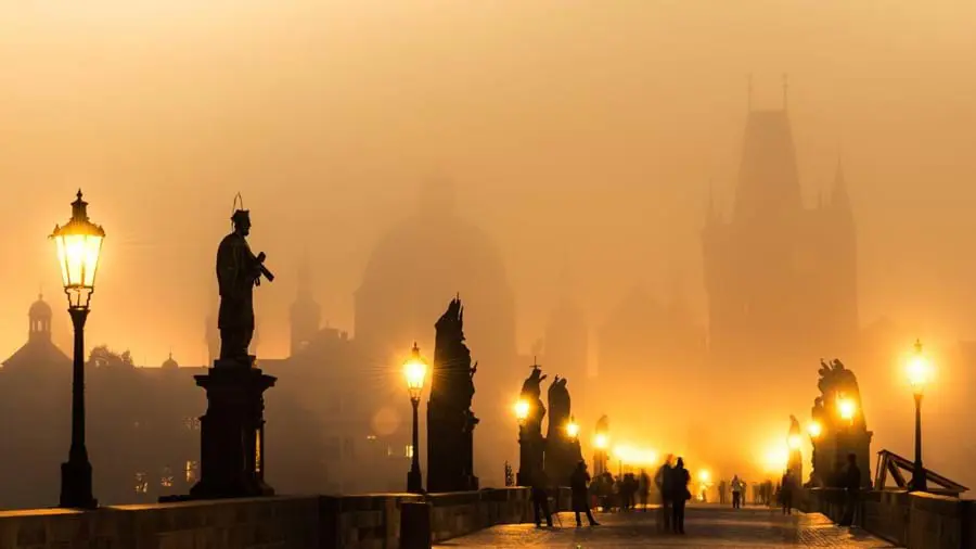 Shot of a bridge lit up by street lamps, with silhouettes of statues lining either side. Silhouettes of buildings in the misty atmosphere at the end 