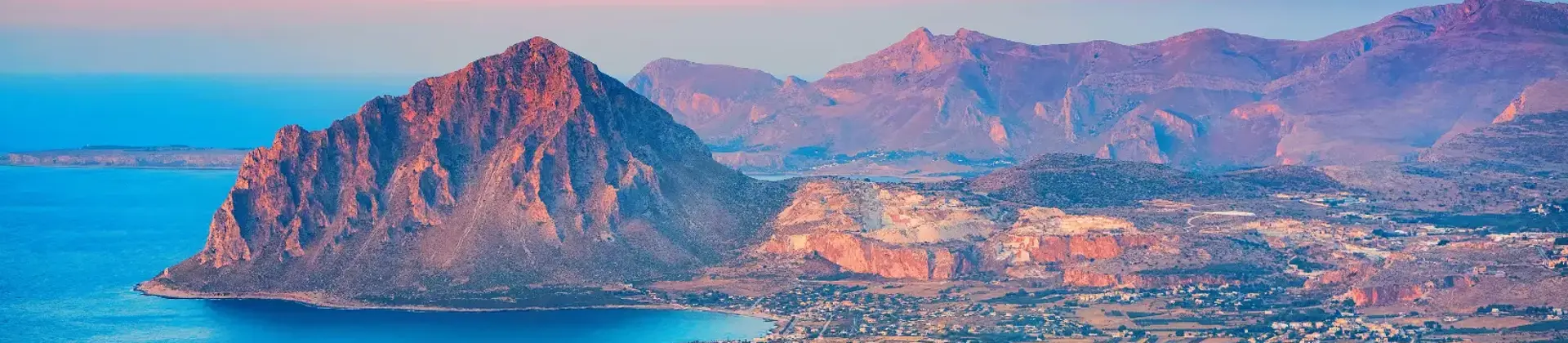 Mountain landscape of Erice, Sicily, showing a rugged hill with green vegetation and buildings clustered near the coastline below