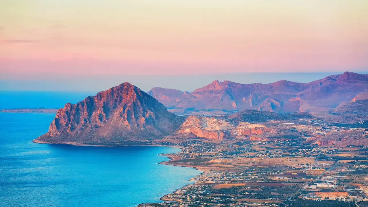 Mountain landscape of Erice, Sicily, showing a rugged hill with green vegetation and buildings clustered near the coastline below