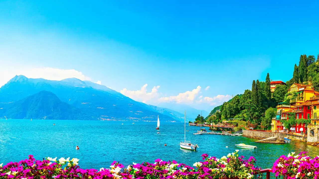 Colourful hillside houses in Lake Como overlooking the lake, where a couple of boats gently float, with mountains in the background