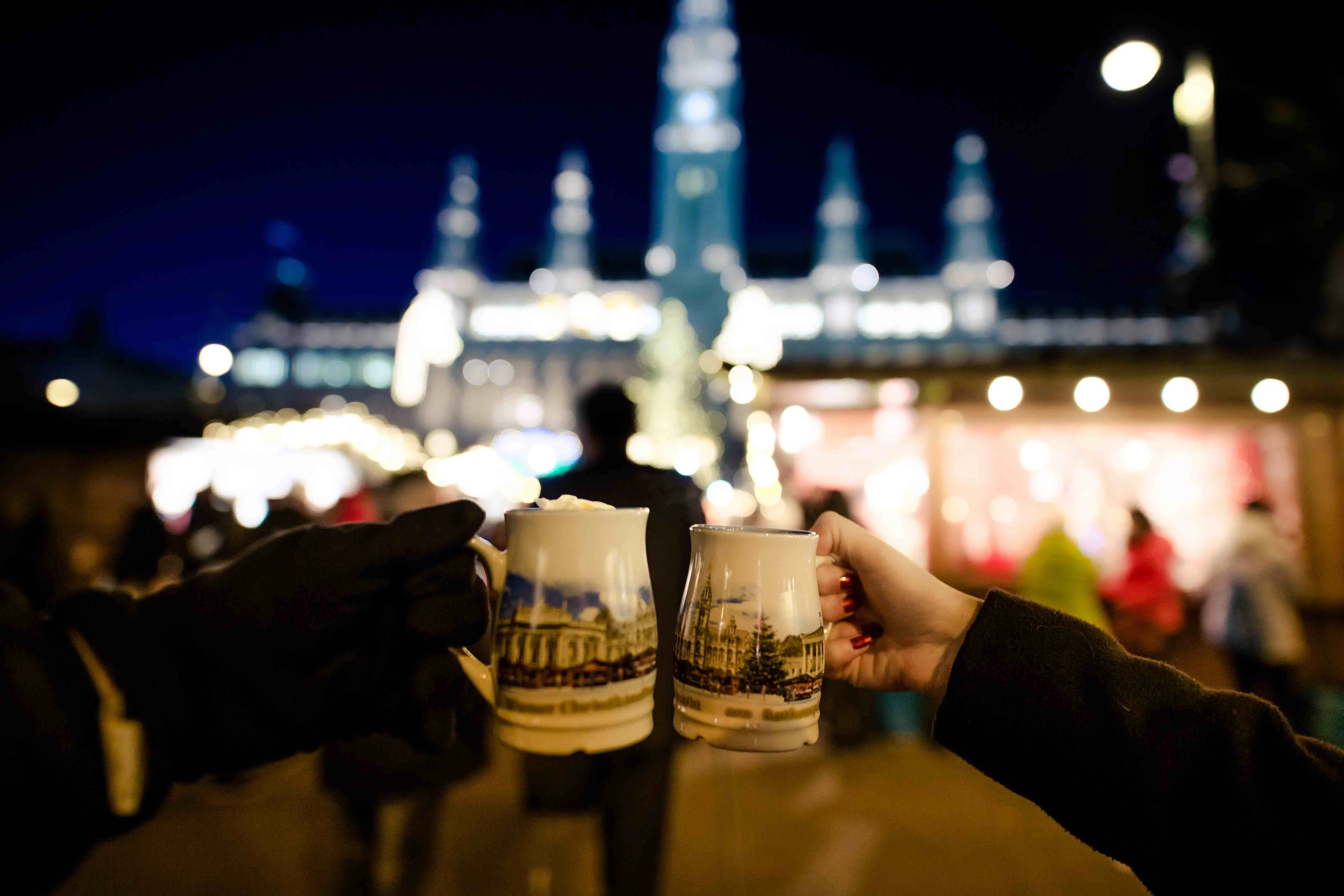 Two arms clinking two mugs of hot chocolate together in front of a blurry, lit up background