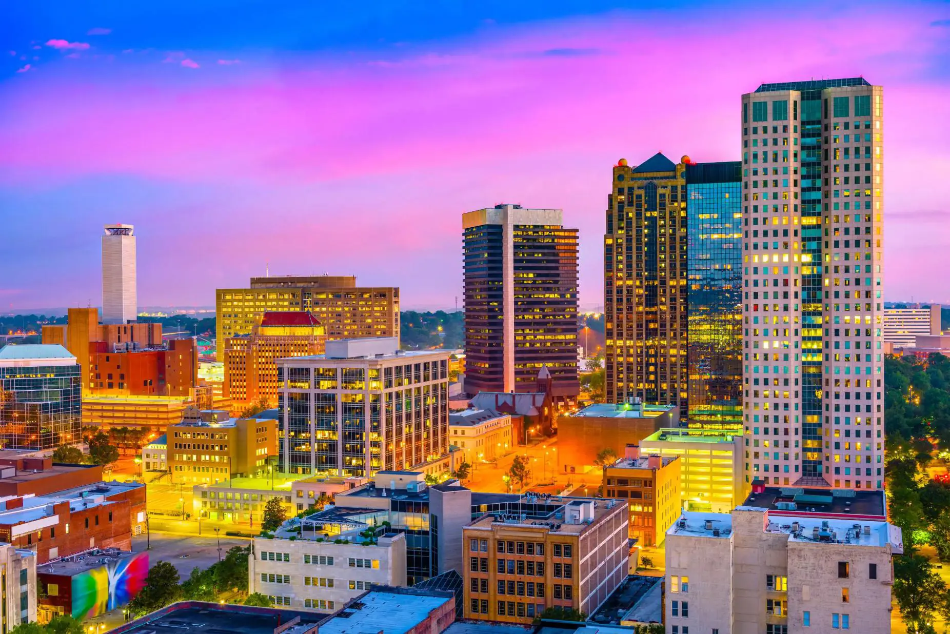 A view of the Birmingham, Alabama skyline at dusk, with skyscrapers and high-rises glowing beneath a vibrant pink and blue sky