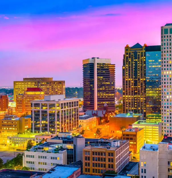 A view of the Birmingham, Alabama skyline at dusk, with skyscrapers and high-rises glowing beneath a vibrant pink and blue sky