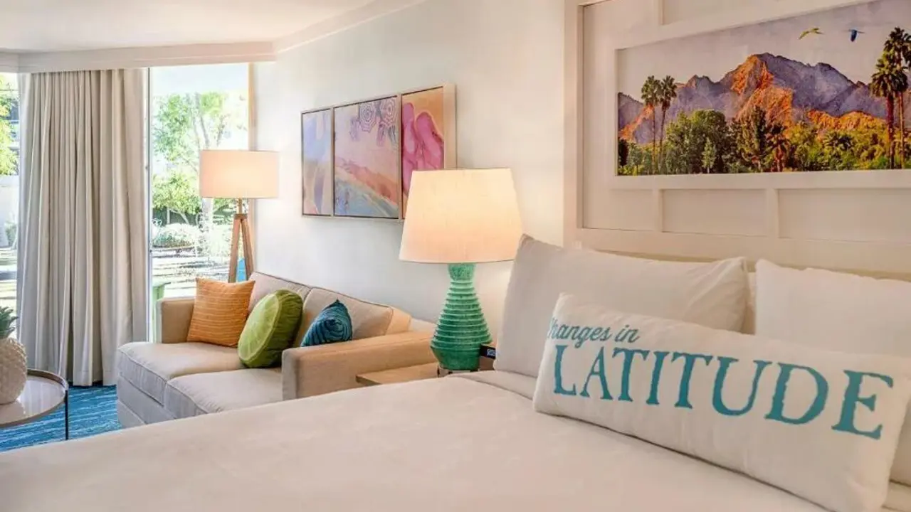 Stylish guest bedroom at the Riviera Resort & Spa, Palm Springs, featuring a large bed with crisp white linens, colourful décor, a seating area, and floor-to-ceiling windows with views of the surrounding landscape