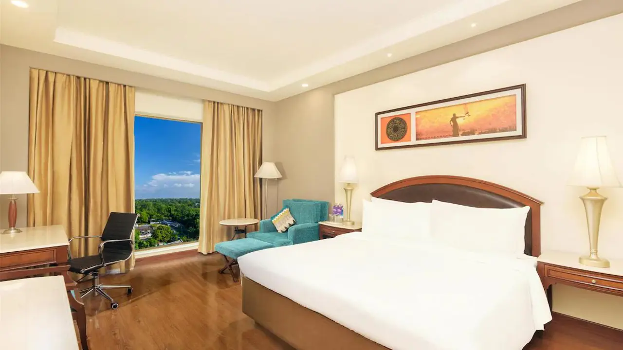 A bright, modern room with a large bed, wooden floors, a teal armchair, and a window overlooking greenery at the Radisson Hotel in Varanasi