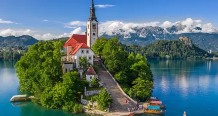 Shot of a church on a small bit of forested land on water, there is a pathway leading up to the church which has some people and boats at the bottom. Behind, there are mountains and clouds, under a blue sky. 