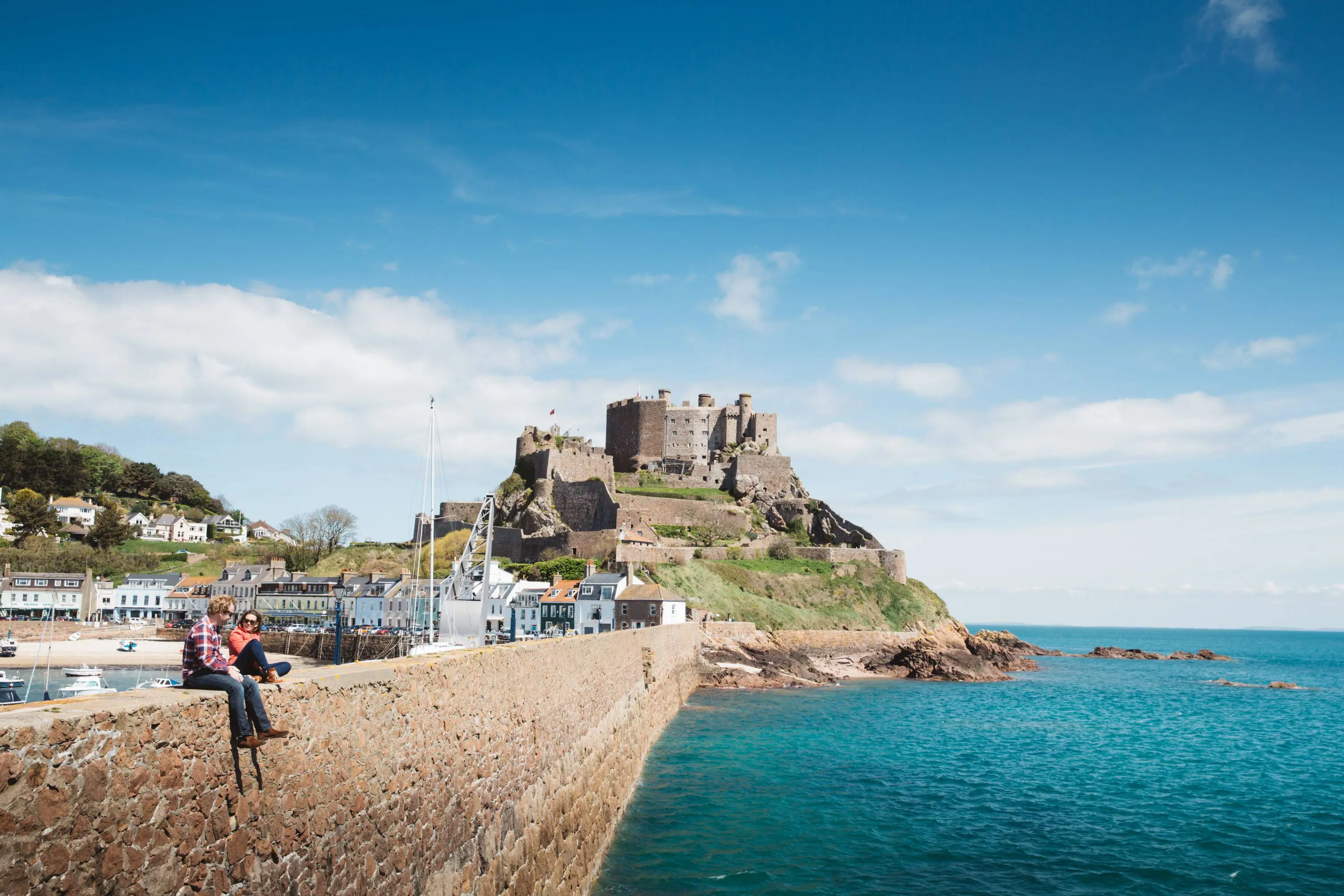 Long shot of Mont Orgueil Castle, showing part of the harbour below it and two people sat on the wall between the harbour and the sea, the buildings beside the harbour and the houses on a hill behind it, on a sunny day with a blue sky 