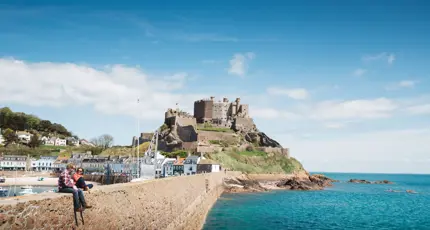 Long shot of Mont Orgueil Castle, showing part of the harbour below it and two people sat on the wall between the harbour and the sea, the buildings beside the harbour and the houses on a hill behind it, on a sunny day with a blue sky 