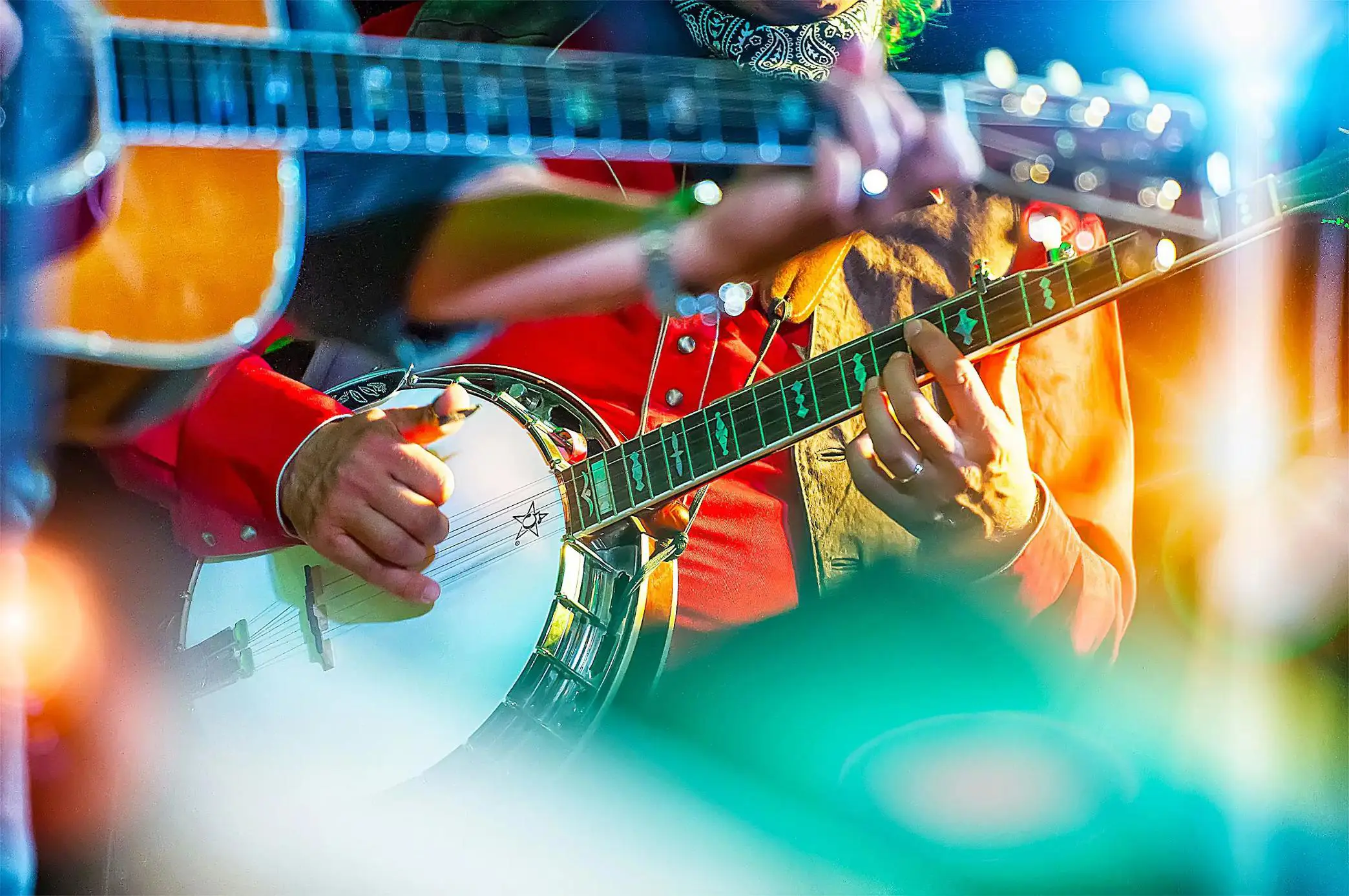 Close-up of a musician playing a banjo on stage, wearing a red shirt, with vibrant stage lighting creating colourful effects in the foreground