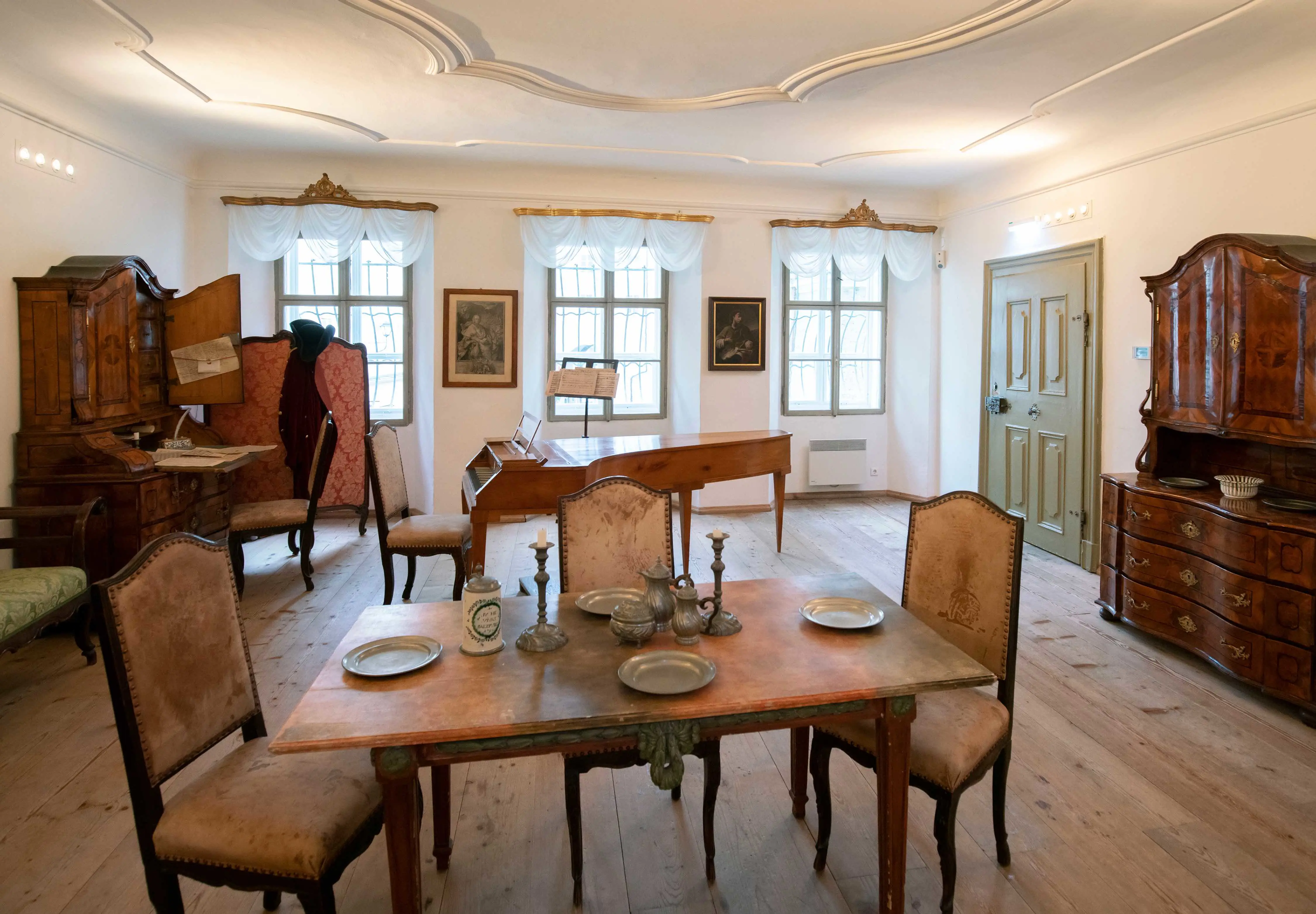 Inside a room with an antique wooden table in the front, with silver plates and two candle holders in the middle, three cream chairs surround the table. Behind is a wooden piano, and another chair at it. A sage door to the right, forwards, a dark wooden cabinet with drawers underneath. To the left, there is another one of these, with the doors open and a desk attached with paperwork on and another chair in front. The back wall has three grid windows and mesh white curtains and paintings between the windows.
