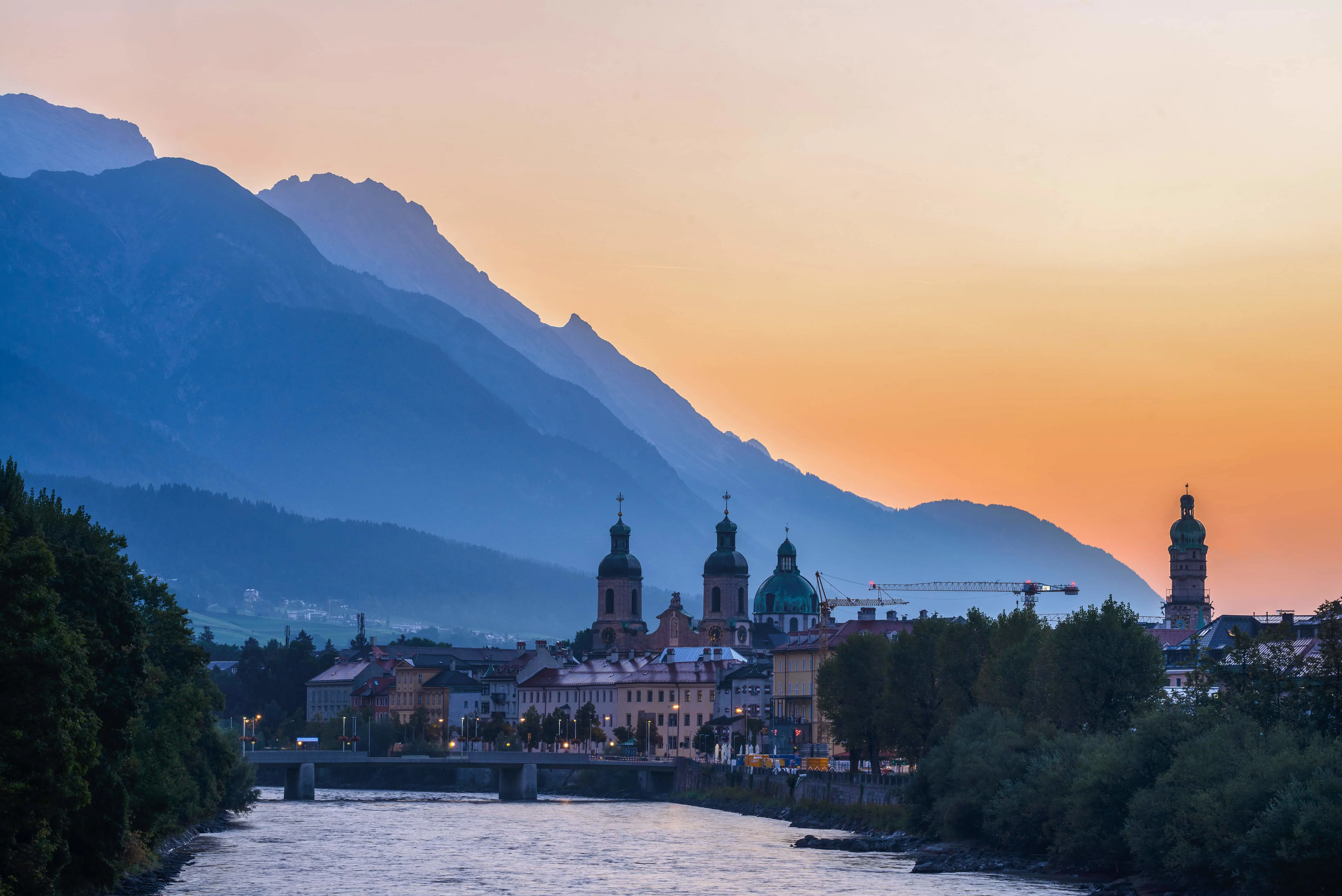 View of the town of Innsbruck at sunset from the river, with a bridge in the forefront and the town's buildings along the waterfront. Two clock towers, one dome shaped building and one tower poke out of the top of the cityscape. The silhouette of a mountain is behind, against an orange sky.