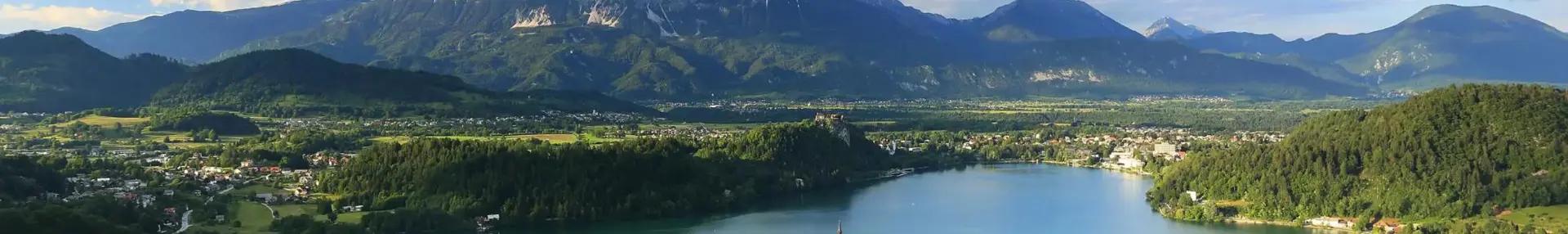 Wide angle shot of Lake Bled, showing its forested surrounding areas, and mountains in the distance. In the middle of the lake, there is a small island with trees and a towered building