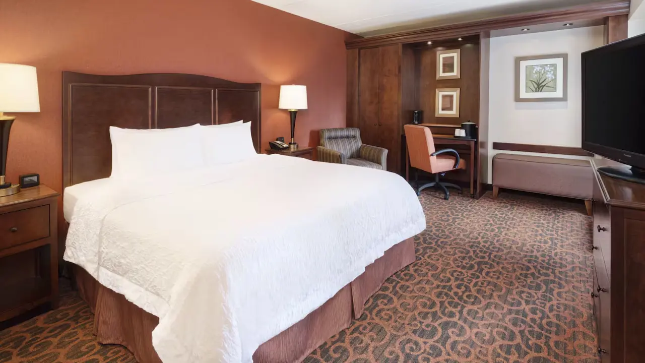 A king room at Hampton Inn Downtown, Cleveland, featuring a spacious king-size bed with crisp white linens, dark wood furnishings, warm-toned décor, and a patterned carpet