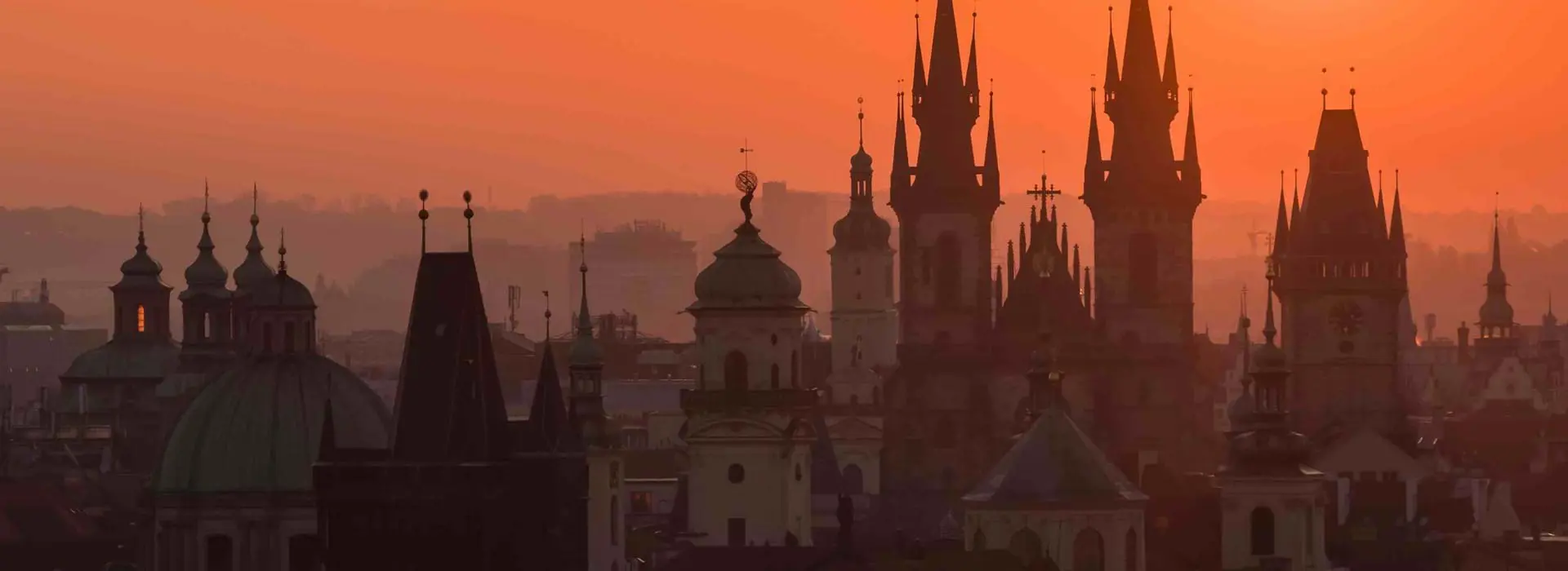 Prague cityscape against an orange sky with silhouettes of tall buildings with spiked turrets